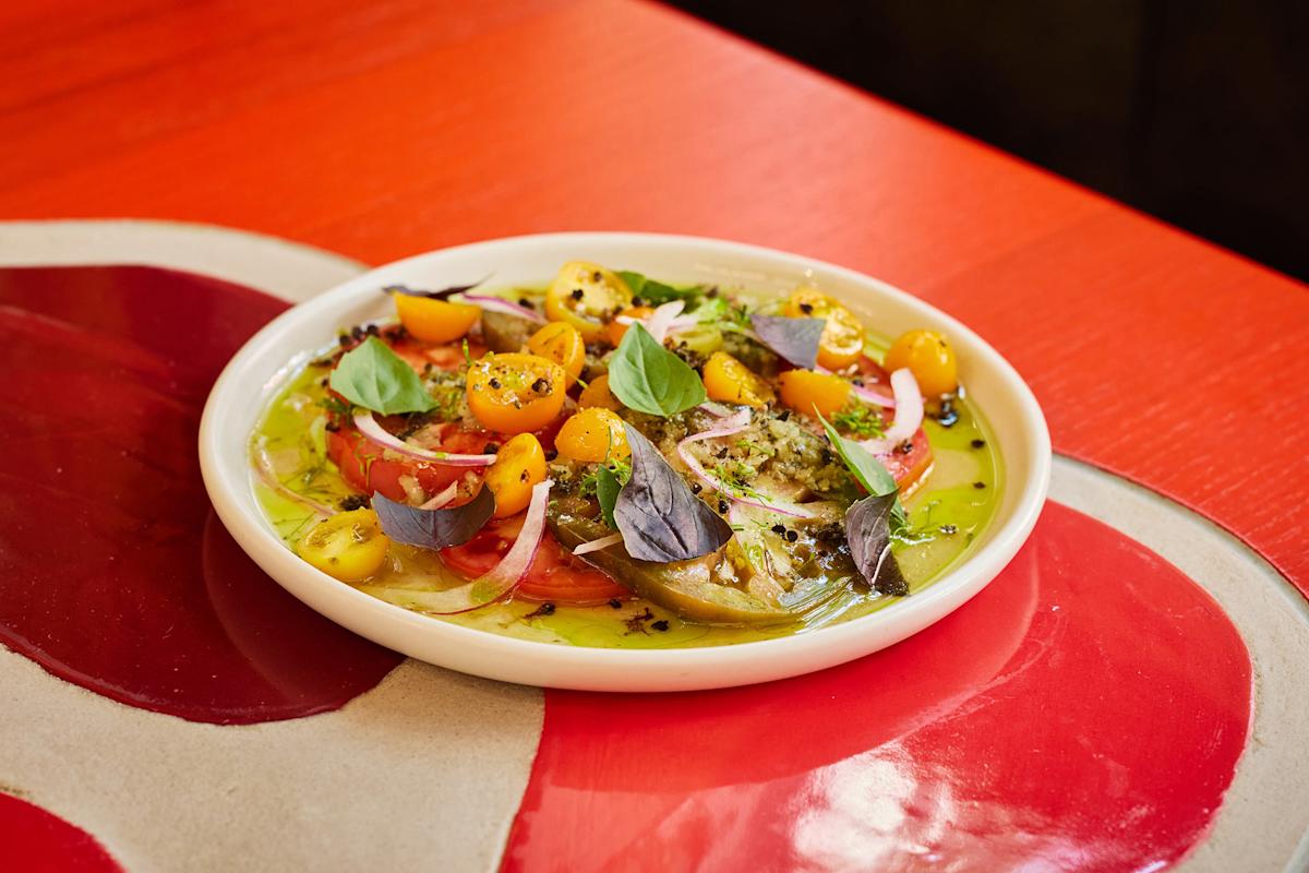 A colorful tomato salad with yellow and red tomatoes, red onion slices, fresh basil leaves, and seasoning, served on a white plate atop a red and tan table.