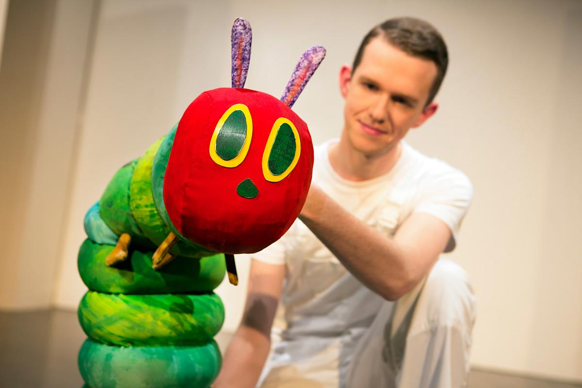 A person in white operates a large, colorful caterpillar puppet with a red head and green body, inspired by “The Very Hungry Caterpillar,” against a plain background.