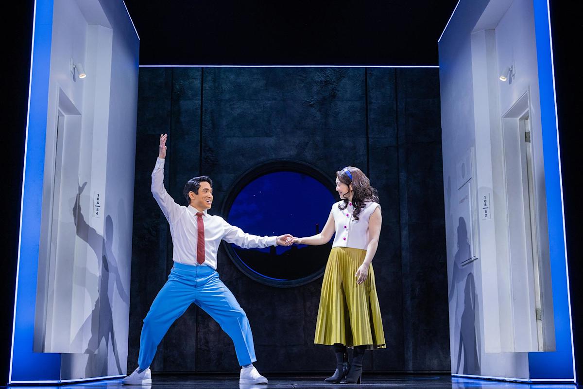 A man in blue pants and a woman in a yellow skirt hold hands on a stage set with white doorframes and a round window, appearing to perform a lively dance.
