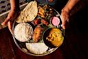 A person holds a round metal plate with rice, papadum, curry, grilled fish, various vegetables, a white crepe, pink dipping sauce, and pickles, all arranged on a banana leaf.