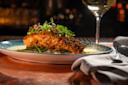 A plated dish featuring a crusted salmon fillet on a bed of greens, topped with microgreens, with a light sauce and a glass of white wine in the background on a softly lit table.