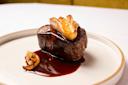 A gourmet dish featuring a piece of steak topped with foie gras, served on a white plate with a rich, dark sauce and garnished with sautéed onions. The presentation is elegant and appealing.