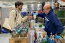 A man in a beige hoodie is receiving a drink from another man wearing blue gloves and a blue hoodie in a store. They are surrounded by various soda siphons and beverage bottles on a counter.
