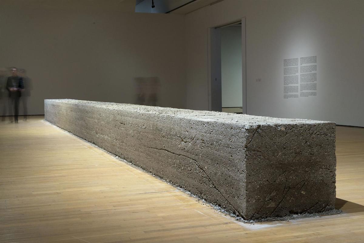 A large, rectangular concrete block sits on a wooden gallery floor, surrounded by plain walls. Two blurred figures stand in the background, and text is displayed on the wall to the right.