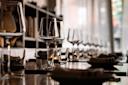 A long dining table set with wine glasses, water glasses, napkins, and small plates in a softly lit restaurant with natural light coming through sheer curtains.