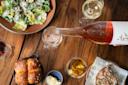 A person pours rosé wine into a glass on a wooden table. Nearby, there are glasses of white and red wine, a plate of Caesar salad, a serving of rolls with butter, and a dish of meat with greens.