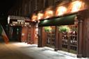 A warmly lit steak and ale house with wooden exterior, green awnings, hanging flower baskets, and an Irish flag on the sidewalk at night. The sign reads “Hunter’s Steak & Ale House.”.