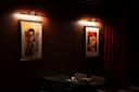 A dimly lit room features two framed vintage-style posters on the wall above a set of chairs and a table. Each poster is individually illuminated by small lights, creating a cozy atmosphere with rich, dark tones.