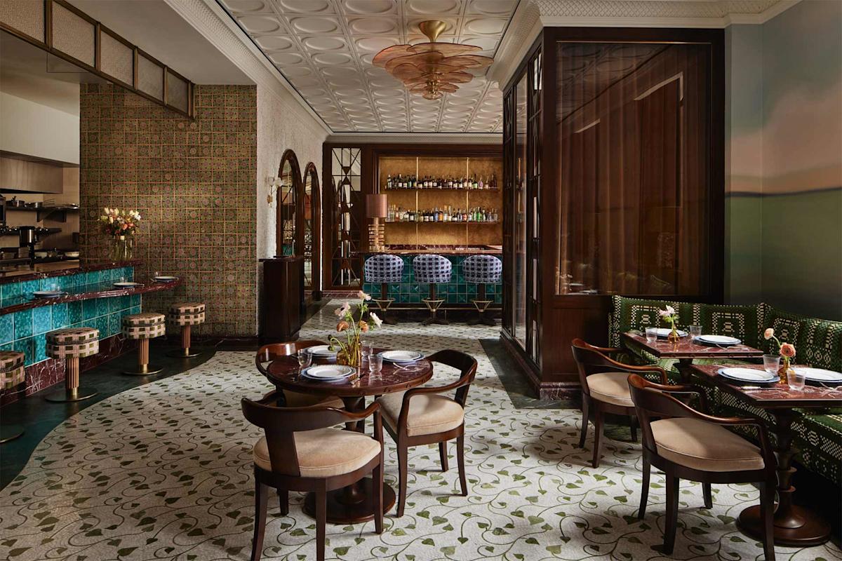 Elegant restaurant interior with patterned floor, wooden tables, cushioned chairs, green banquette seating, a teal-tiled bar, bar stools, and decorative lighting. Shelves with bottles are visible behind the bar area.