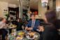 People are dining at a stylish restaurant. A man in a suit and a woman are at a table with dishes and drinks.