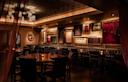 A dimly lit, elegant restaurant with dark leather seating, wooden tables, and eclectic framed art on the walls. Soft warm lighting creates a cozy, intimate atmosphere.