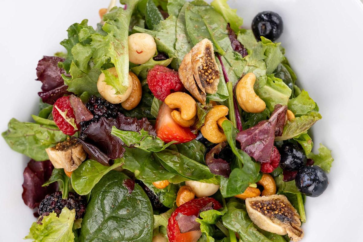 A close-up of a salad with leafy greens, cashews, dried figs, strawberries, blackberries, raspberries, and blueberries in a white bowl.