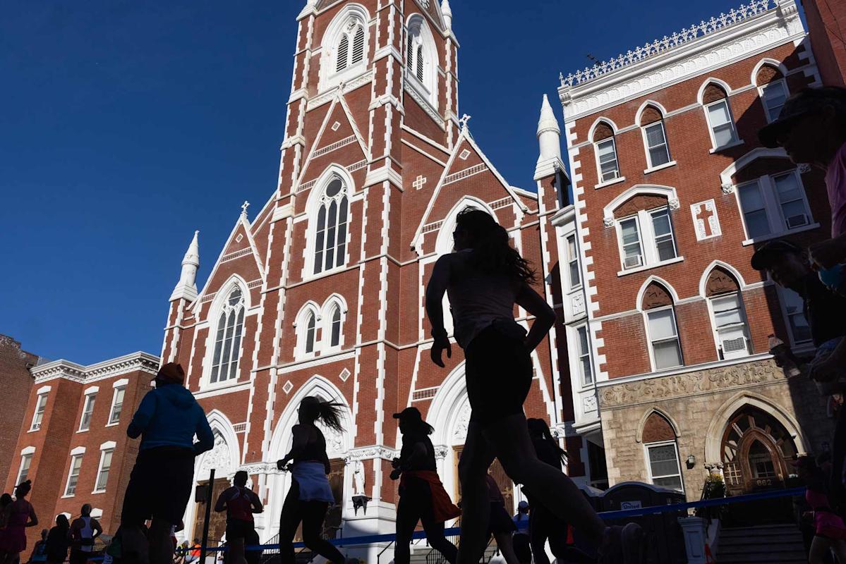 Silhouetted runners pass in front of a large brick church with white trim, featuring a tall steeple. The sky is clear, highlighting the building's gothic architectural style.