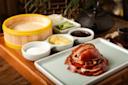 Slices of roasted duck are served on a rectangular plate. Accompaniments include hoisin sauce, sliced green onions, cucumbers, sugar, and yellow pickles. A bamboo steamer contains pancakes, and a teapot is in the background.