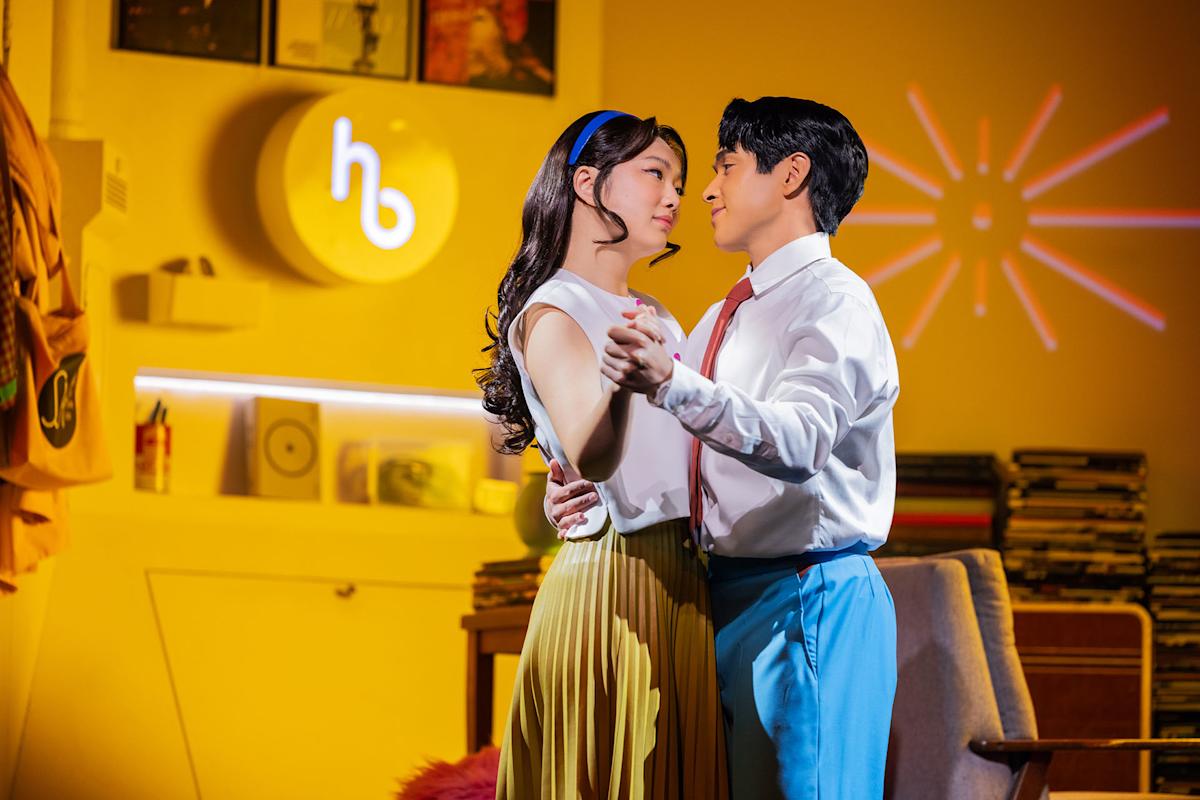 A man and woman dance closely in a warmly lit, retro-styled room with yellow walls, records, and vintage decor in the background. Both are smiling and gazing into each other's eyes.
