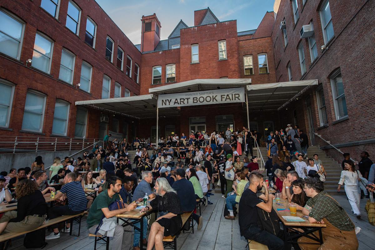 A large crowd sits at tables and on steps outside a brick building with a banner reading “NY Art Book Fair.” People are socializing, eating, and drinking in a lively outdoor atmosphere.
