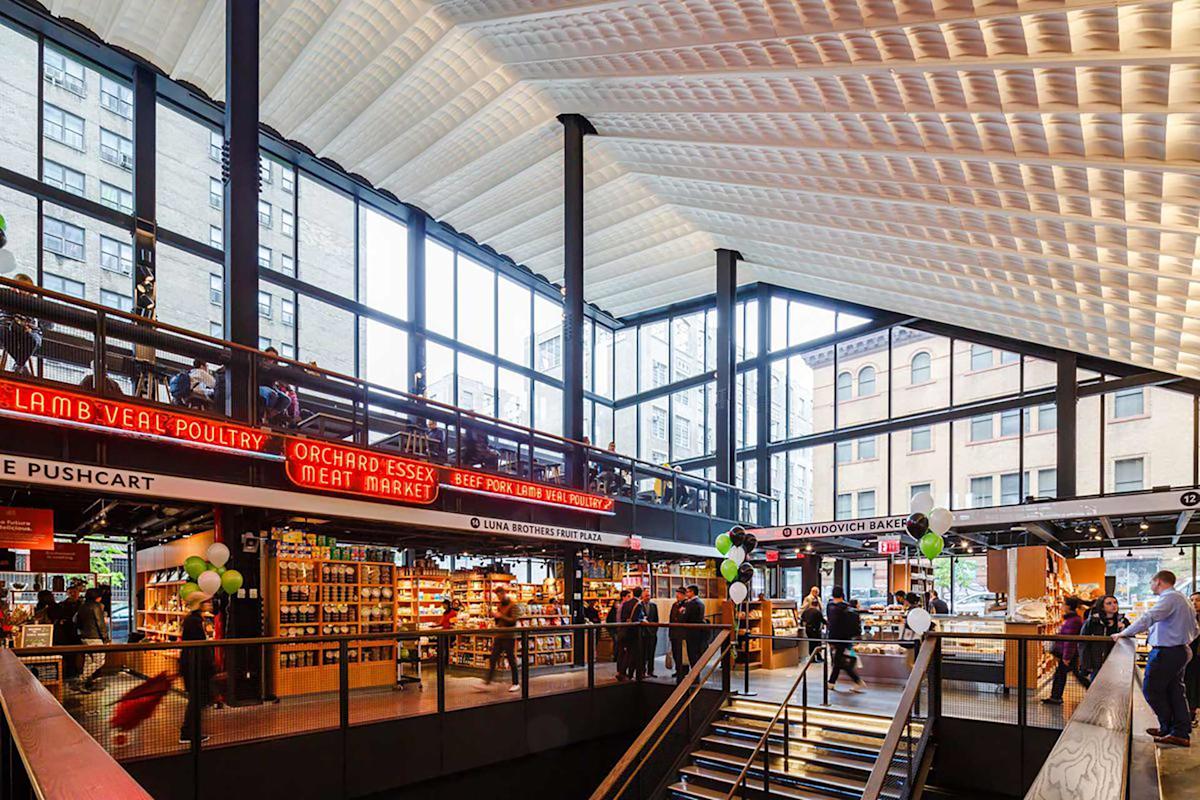 A modern indoor market with large windows, high patterned ceiling, bright lighting, and various vendors, including a meat market and bakery. Shoppers and staff are visible, and green-white balloons decorate the space.