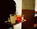 A person carries a tray with a glass of yellow drink, a red drink with a lemon slice, a clear drink, a glass of water, and a green bottle in a dimly lit indoor setting.