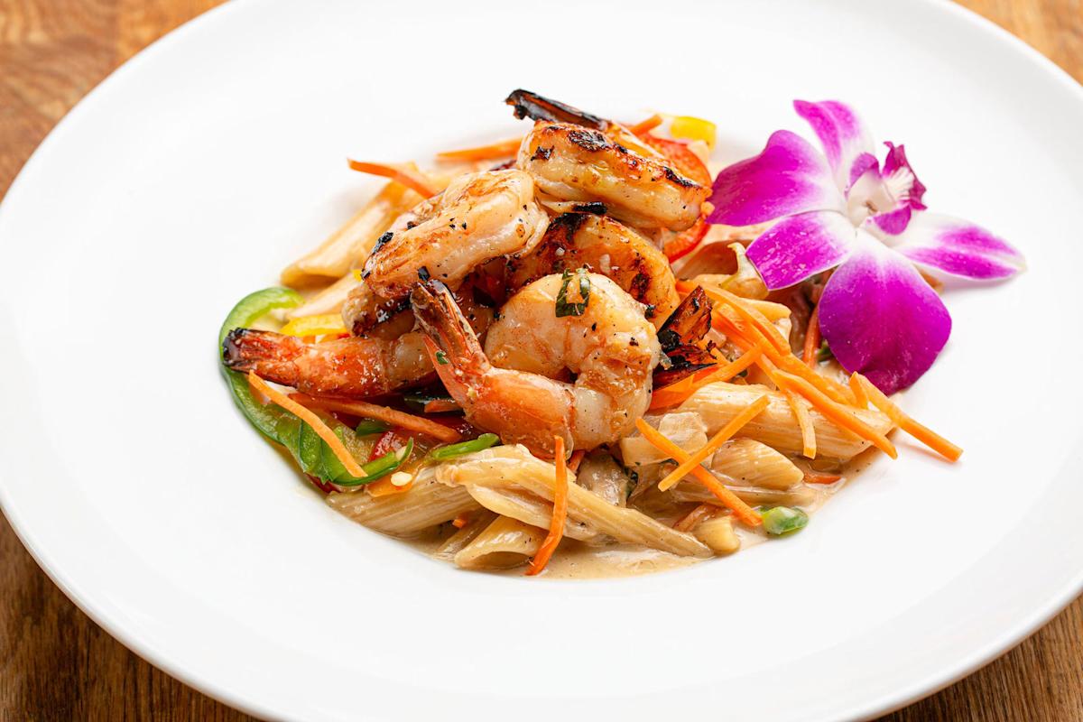 Grilled shrimp served on penne pasta with julienned carrots and snap peas in a creamy sauce, garnished with a vibrant purple orchid on a white plate.