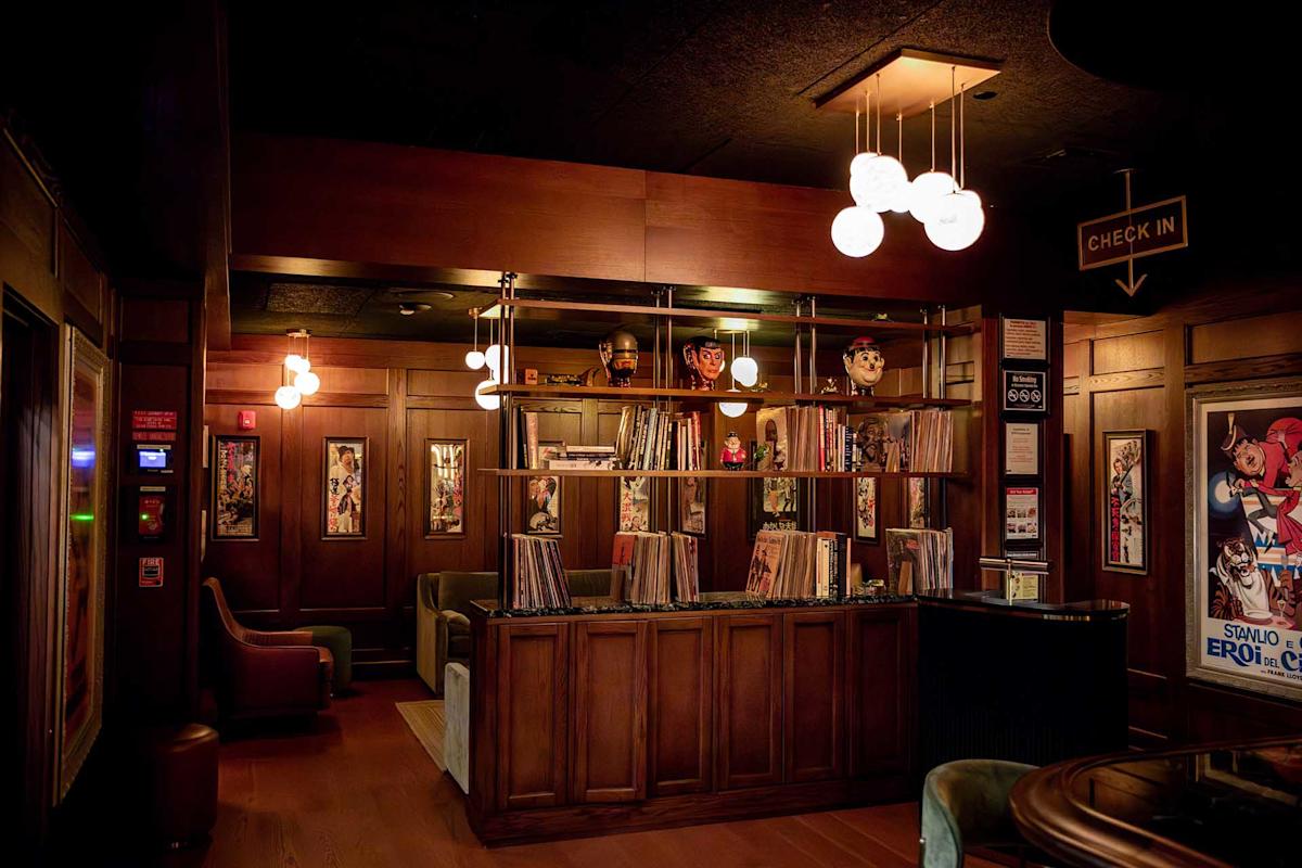A cozy, dimly lit lounge with wood paneling, vinyl records on shelves, vintage posters, plush chairs, and a “Check In” sign hanging from the ceiling. Warm lighting creates a welcoming atmosphere.