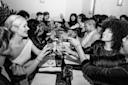 A group of people sit around a long table at a restaurant, raising their glasses in a celebratory toast. They are smiling and laughing, creating a joyful, festive atmosphere. The image is in black and white.
