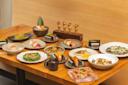 A wooden table covered with a variety of Japanese dishes, including sushi, sashimi, grilled fish, small appetizers, and desserts, all artfully plated and arranged in a modern dining setting.