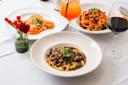 Three plates of Italian pasta dishes and two glasses of drinks on a white tablecloth, with a small vase of red flowers. One pasta has mushrooms, another has red sauce, and the third is topped with herbs.