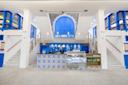A bright, modern café interior features blue and white tile patterns, a counter with a glass display case, two staircases ascending on either side, and vibrant blue accents on windows and doors.