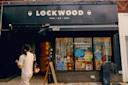 A person walks past the storefront of LOCKWOOD, a shop with a black sign reading "home | gift | paper." The window displays colorful decorations and various items for sale inside the store.