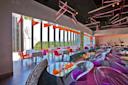 A modern restaurant interior with large windows overlooking a city skyline and greenery. Tables are set with white cloths, surrounded by colorful chairs. Neon geometric lights hang from the ceiling, adding a vibrant touch to the space.