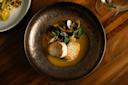 A dish featuring seared scallops on a rich orange sauce, garnished with clams, zucchini, and herbs. It's elegantly presented in a textured dark bowl on a wooden table, with a partial view of another dish and a wine glass beside it.