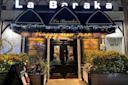 Front of La Baraka restaurant decorated with "Happy New Year" banner and lights. Two stained glass windows flank the entrance. An "A" rating and "Open" sign are on the door. Plants are on either side of the entrance. Nighttime setting.