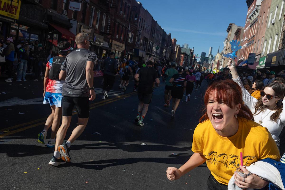 A woman in a yellow shirt cheers with excitement among a crowd of runners. Other spectators are visible, and rows of buildings line both sides of the sunny street. Confetti is scattered on the ground.