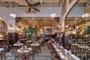 Spacious restaurant interior with high ceilings, white subway tile walls, and elegant lighting. Wooden tables and chairs are neatly arranged, each set with glassware and white napkins. Large potted plants add greenery to the warm atmosphere.