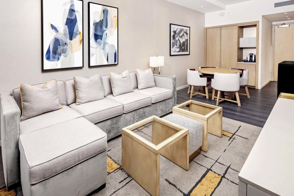 A modern living room with a large gray sectional sofa, two wooden coffee tables, and abstract art on the wall. A small dining area with a round table and four chairs is in the corner, next to a kitchenette. The room has a contemporary decor.