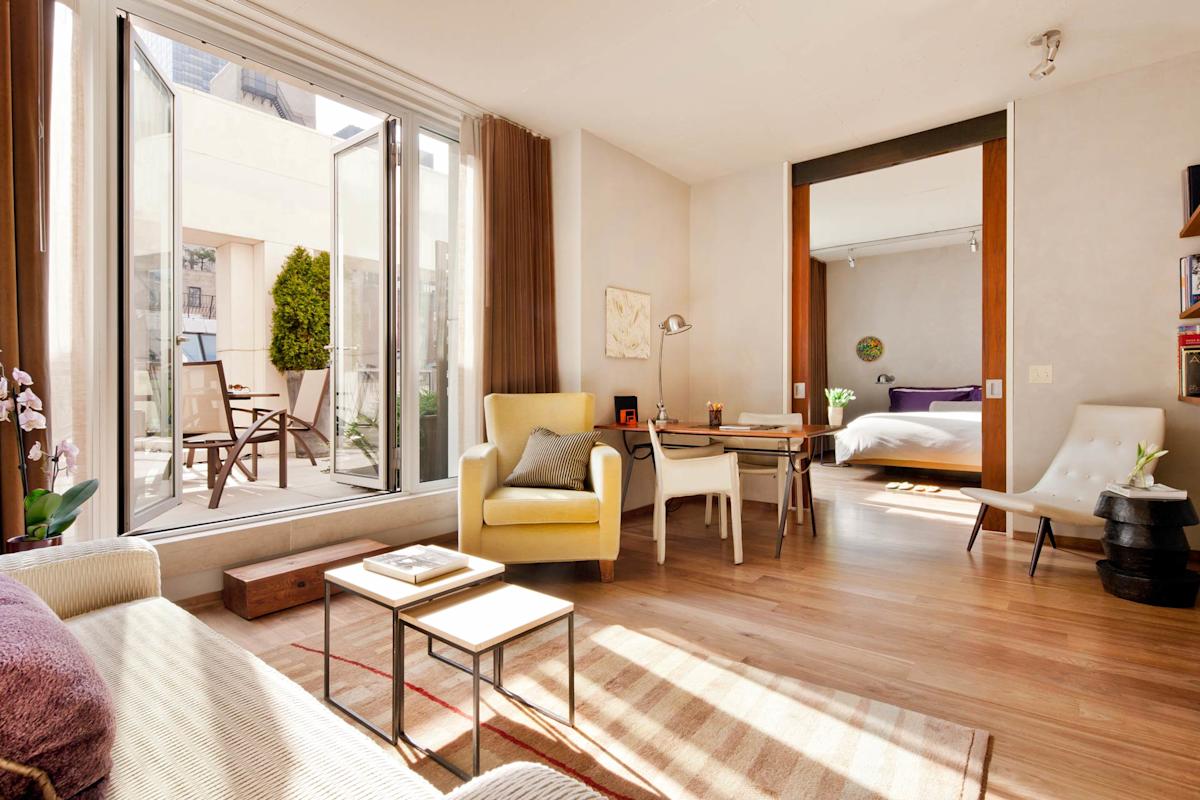 Bright, modern apartment with wood floors, open living area, desk, yellow armchair, and sliding doors leading to a bedroom. Large glass doors open to a sunny patio with outdoor furniture.