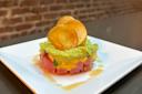 A plated dish featuring a circular stack of diced raw fish, likely tuna, layered with avocado and garnished with crispy potato chips on top. The presentation is simple, set against a white plate with a brick wall in the background.