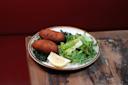 A plate with two golden-brown croquettes, a fresh green salad, and a lemon wedge, set on a rustic wooden table with a red background.