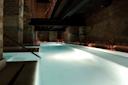 A long indoor pool with glowing white water, surrounded by exposed brick walls and subtle lantern lighting, creating a calm and atmospheric setting under a metal walkway.