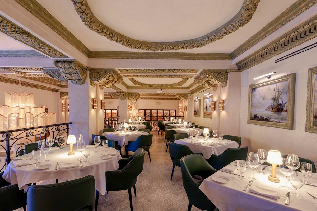 Elegant restaurant interior with ornate ceiling and intricate molding. Tables are set with white tablecloths, wine glasses, and lamps. Green chairs surround each table. Warm lighting complements the classic decor and framed wall art.