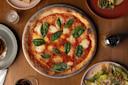 A freshly baked pizza topped with tomato sauce, melted mozzarella, and basil leaves on a wooden table. Surrounding the pizza are plates with food, a lit candle, a pepper shaker, and a glass of red wine.
