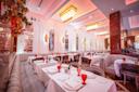 Elegant restaurant interior with white tablecloths, red glassware, plush gray seating, large mirrors, arched wall decor, chandeliers, and warm ambient lighting. Ornamental flowers decorate one side of the room.