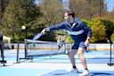 A man in athletic wear plays pickleball on an outdoor court, reaching forward with a paddle to hit a ball. Trees and people are visible in the background.