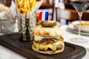 A cheeseburger topped with caramelized onions, melted cheese, and a pickle, served on a wooden board with a small French flag. A metal cup with French fries and a glass of red wine are in the background.