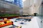 Modern outdoor patio with cushioned seating, tables, and chairs on a rooftop terrace surrounded by tall glass and stone buildings, with greenery along the edges and a partly cloudy sky above.