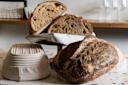 Sliced artisanal bread with a rustic crust is displayed on a round wooden stand, partially covered by a white and green cloth. Two textured proofing baskets are visible on a speckled countertop, with empty wine glasses in the background.