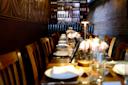 A long wooden dining table set for a meal with white plates, glasses, and floral centerpieces. The restaurant has dim lighting, a brick wall on the right, and a shelf filled with wine bottles in the background.