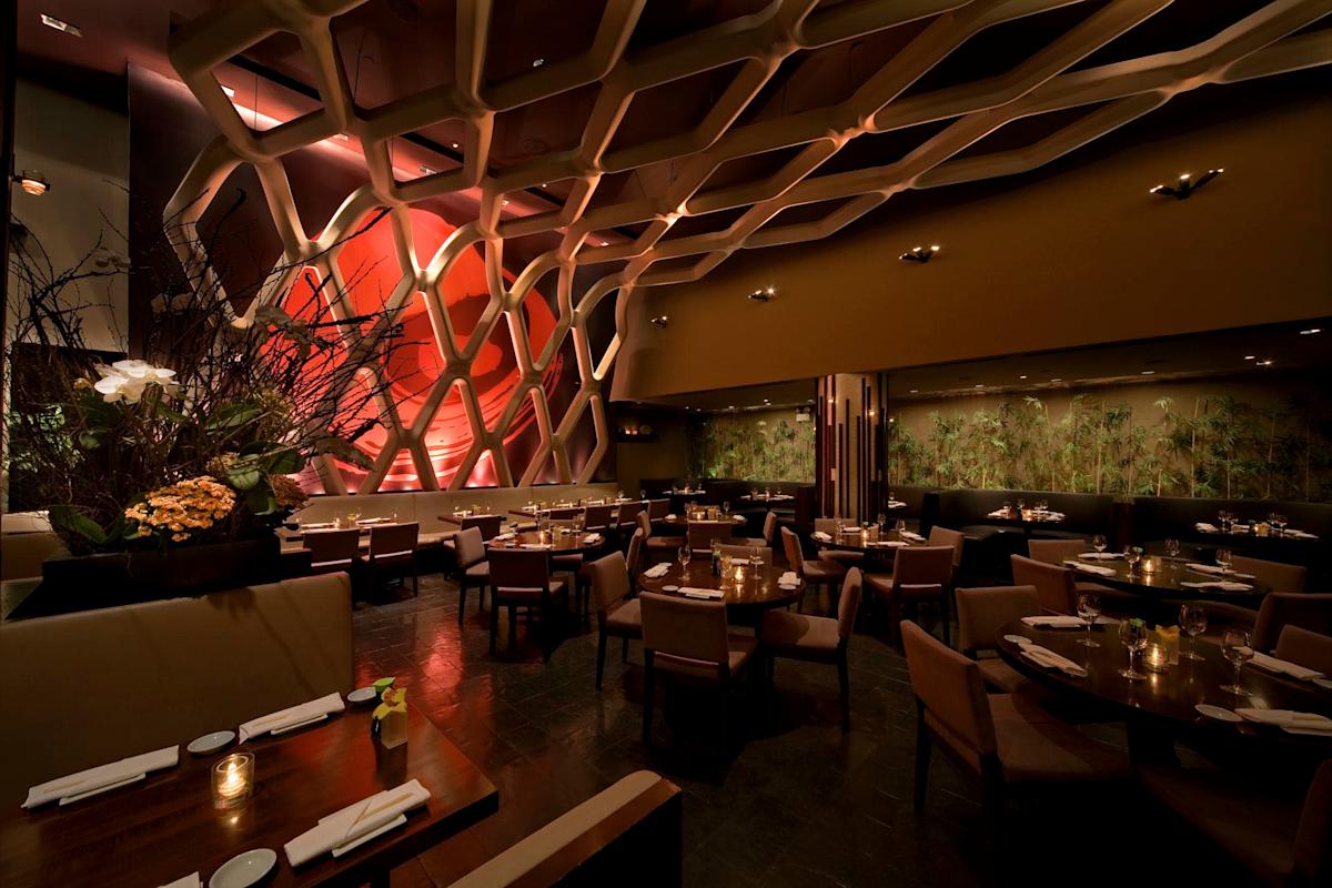 Dimly lit, elegant restaurant interior featuring contemporary decor with a geometric wall design, round wooden tables set with white napkins, and cushioned chairs. Floral arrangements enhance the ambiance.