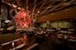 Dimly lit, elegant restaurant interior featuring contemporary decor with a geometric wall design, round wooden tables set with white napkins, and cushioned chairs. Floral arrangements enhance the ambiance.