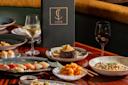 A table set with gourmet dishes including sushi, steak, pasta, and shrimp, accompanied by glasses of white and red wine, with a dark Catch menu upright in the center.